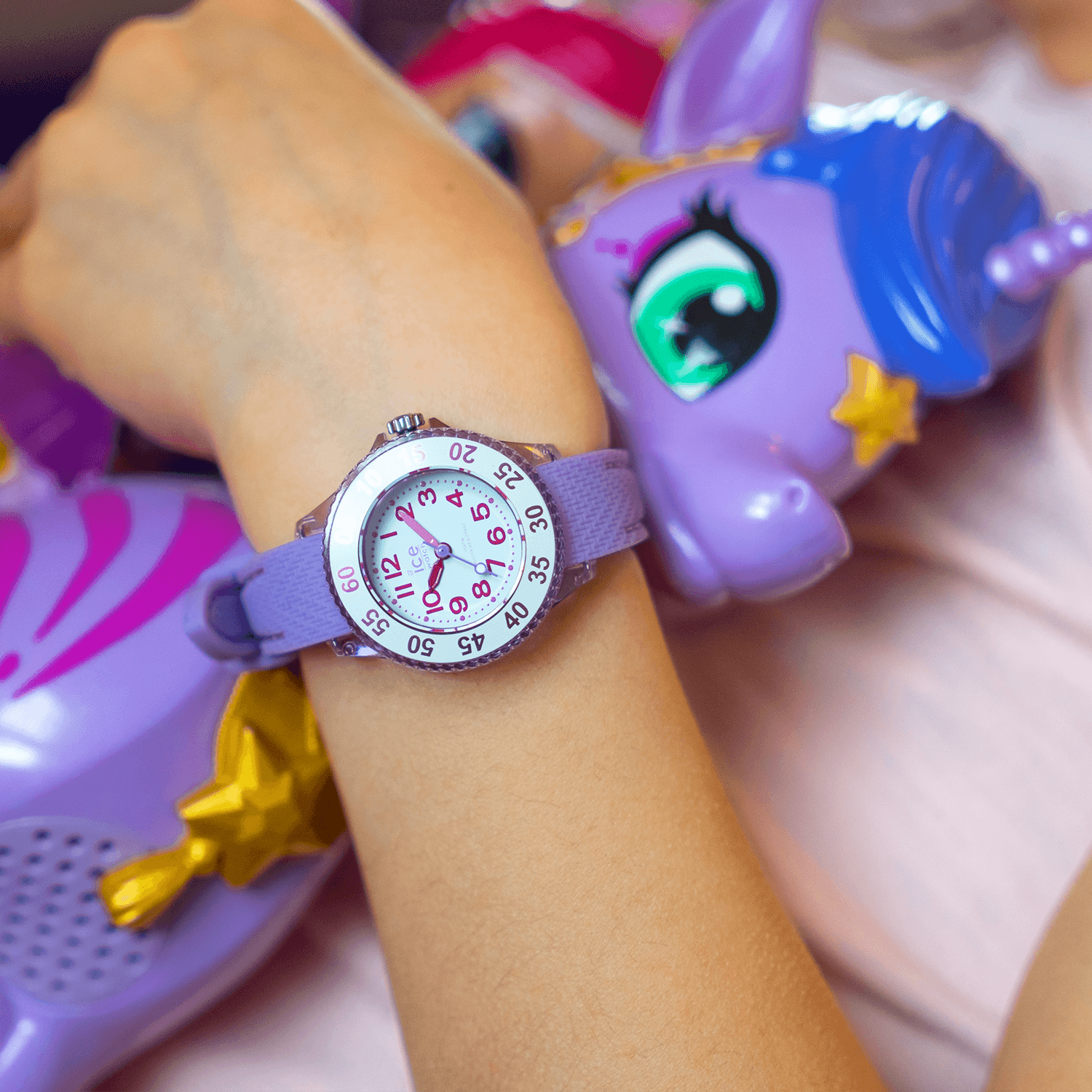 Kinderarm trägt eine lila Armbanduhr mit gut lesbarem Zifferblatt neben bunten Einhorn-Spielzeugen in passenden Farbtönen