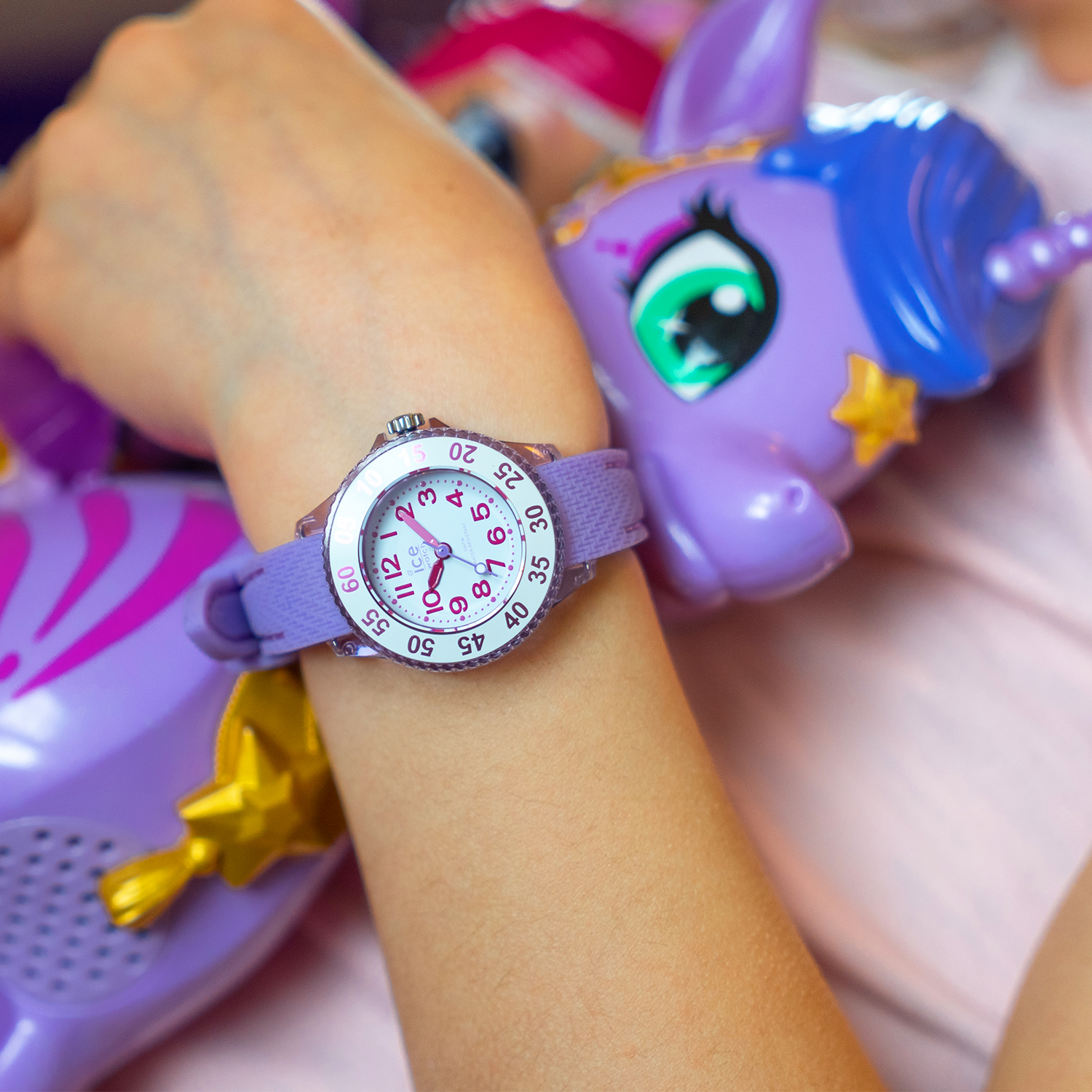 Kinderarm trägt eine lila Armbanduhr mit gut lesbarem Zifferblatt neben bunten Einhorn-Spielzeugen in passenden Farbtönen