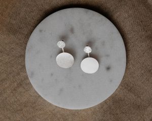 Silberfarbene Ohrstecker mit kleiner strukturierter Scheibe und größerem runden Anhänger in matter Optik, dekorativ auf einer runden Marmorplatte präsentiert.