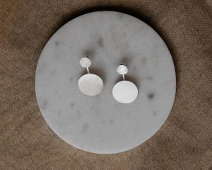 Silberfarbene Ohrstecker mit kleiner strukturierter Scheibe und größerem runden Anhänger in matter Optik, dekorativ auf einer runden Marmorplatte präsentiert.