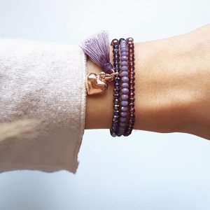 Mehrreihiges lila Perlenarmband mit Quaste und roségoldfarbenem Herzanhänger am Handgelenk einer Person.