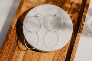 Silberfarbene Ohrstecker mit zwei verbundenen ovalen Ringen in matter Optik, dekorativ auf einer runden Marmorplatte in einer Holzschale präsentiert.