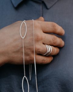 Hand trägt eine lange silberne Halskette mit zwei ovalen Anhängern und schmalem Stabelement sowie mehrere silberne Ringe.