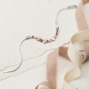 Silbernes Armband mit Herz-, Figuren-, Zahlen- und funkelnden Schmuckelementen auf hellem Untergrund neben einem Stoffband.