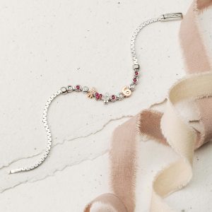 Silbernes Armband mit Herz-, Figuren-, Zahlen- und funkelnden Schmuckelementen auf hellem Untergrund neben einem Stoffband.