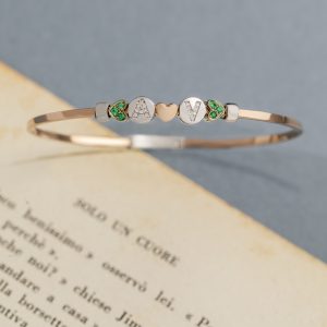 Roségoldfarbener Armreif mit LOVE-Buchstaben, Herzmotiv und grünen Schmucksteinen vor einer geöffneten Buchseite.