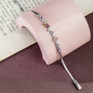 Feines silbernes Armband mit bunten Schmuckelementen, Herz-, Figuren- und Buchstabenmotiven auf rosa Schmuckhalter vor Buchseite.