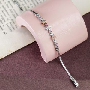 Feines silbernes Armband mit bunten Schmuckelementen, Herz-, Figuren- und Buchstabenmotiven auf rosa Schmuckhalter vor Buchseite.