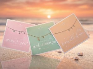 Drei feine Armbänder mit kleinen Anhängern liegen auf pastellfarbenen Spruchkarten vor einem Strandhintergrund bei Sonnenuntergang.