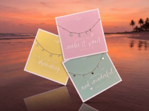 Drei feine Armbänder mit kleinen Anhängern liegen auf pastellfarbenen Spruchkarten vor einem Strandhintergrund bei Sonnenuntergang.
