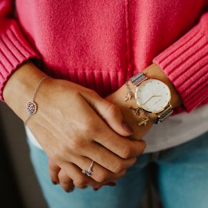 Nahaufnahme einer Frau, die eine roségoldfarbene Armbanduhr mit dekorativem Zifferblatt, ein Charm-Armband, ein silbernes Armband und einen Ring trägt, kombiniert mit einem leuchtend pinkfarbenen Pullover und hellblauen Jeans.