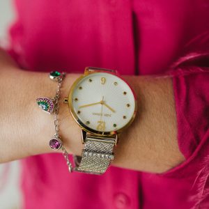 Nahaufnahme einer goldfarbenen Damenarmbanduhr mit weißem Zifferblatt und Armband, kombiniert mit einem silbernen Armband mit bunten Edelsteinanhängern und einer leuchtend pinkfarbenen Bluse.