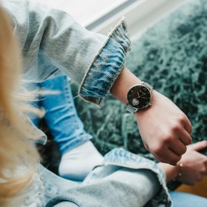 Person trägt eine silberne Mesh-Armbanduhr mit schwarzem Zifferblatt und herzförmigem Zierdetail, kombiniert mit einer hellen Jeansjacke und Jeans in einem lässigen Umfeld.