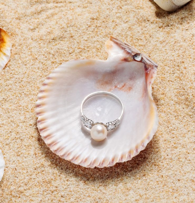 Perlenring auf einer Muschel im Sand, umgeben von Muscheln – Symbol für sommerlichen Schmuck und stilvolle Strandlooks.