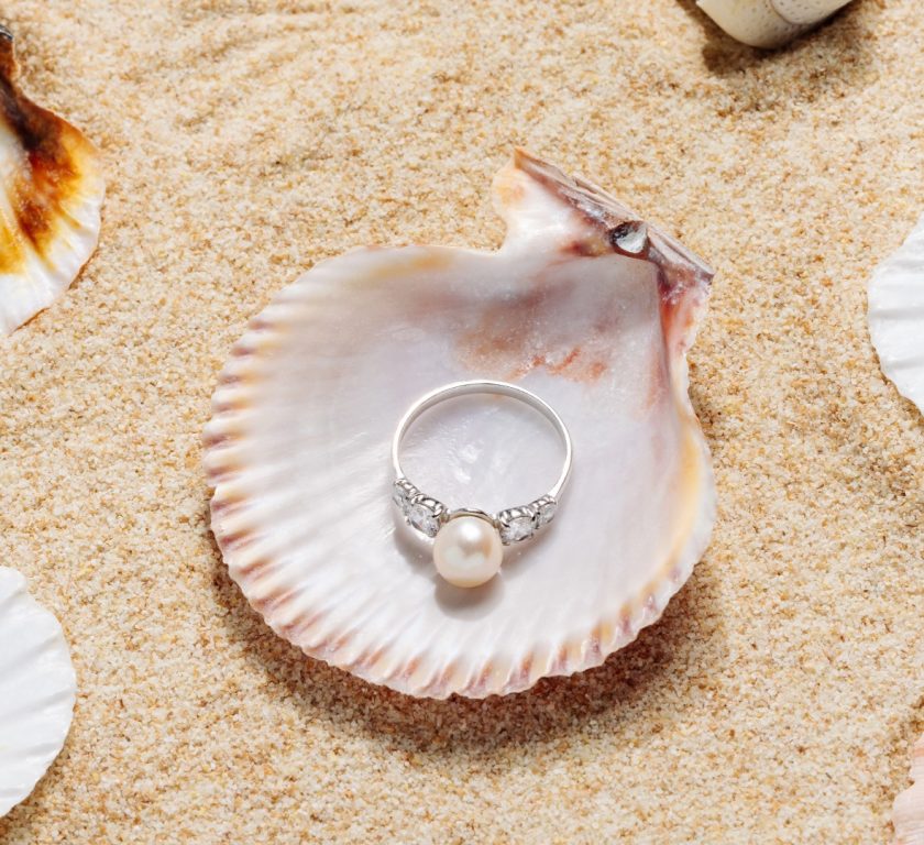 Perlenring auf einer Muschel im Sand, umgeben von Muscheln – Symbol für sommerlichen Schmuck und stilvolle Strandlooks.
